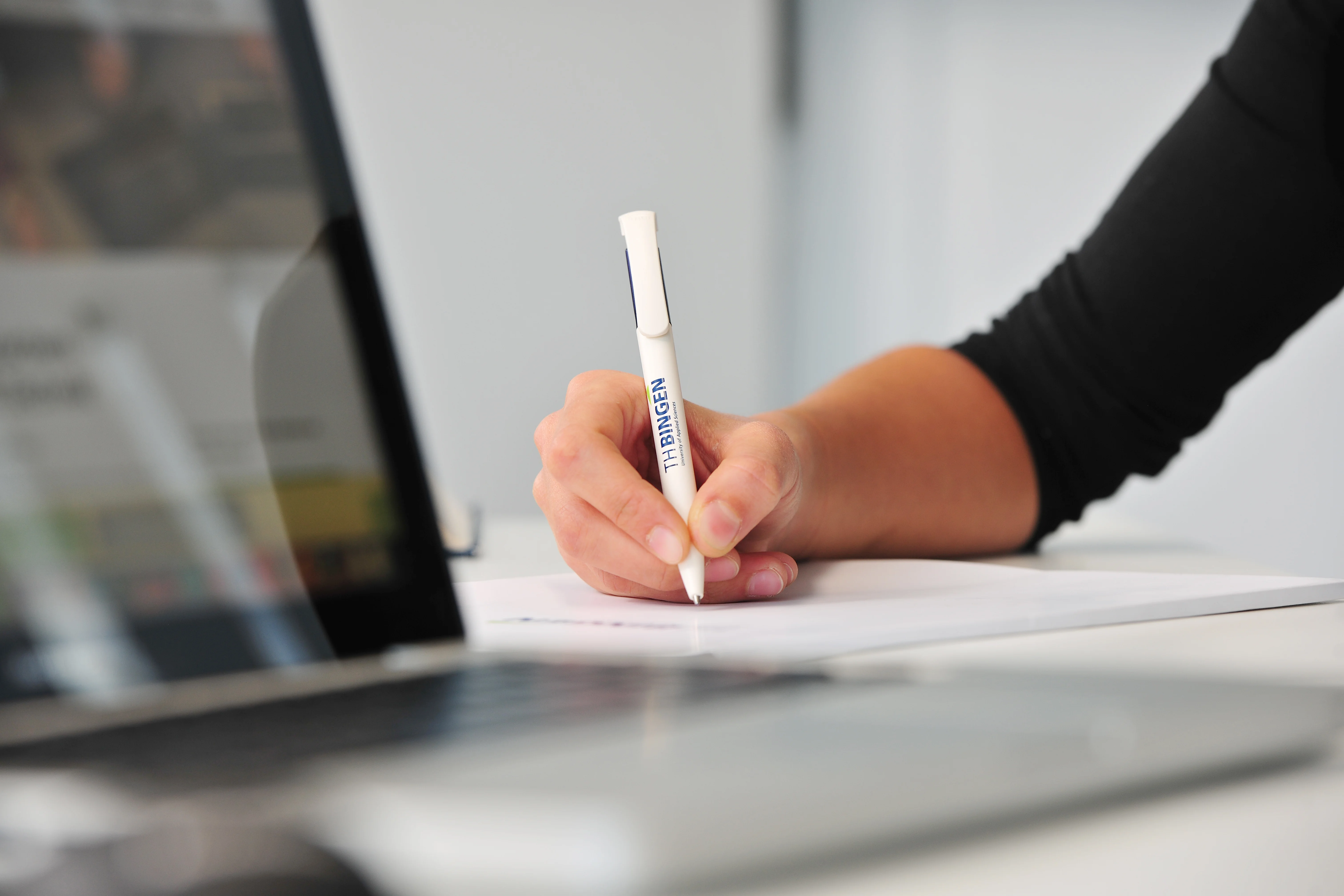 Auf dem Bild ist eine Hand zu sehen, die einen TH Bingen Kuli in der Hand hält und ein Laptop im Vordergrund verschwommen. 
