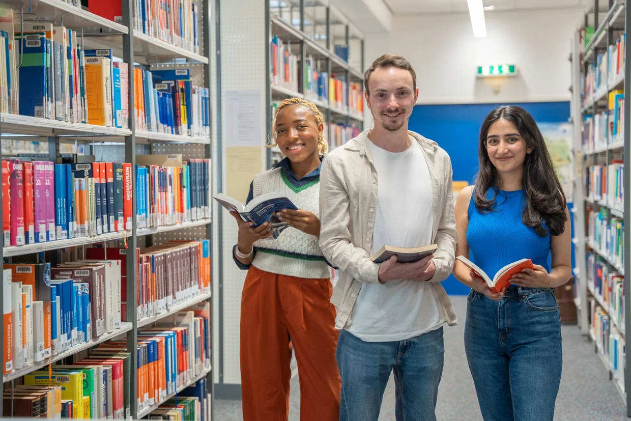 Schwarze Studentin, weißer Student und weiße Studentin zwischen zwei Buchreihen in der Bibliothek halten Bücher in den Händen und lächeln in die Kamera