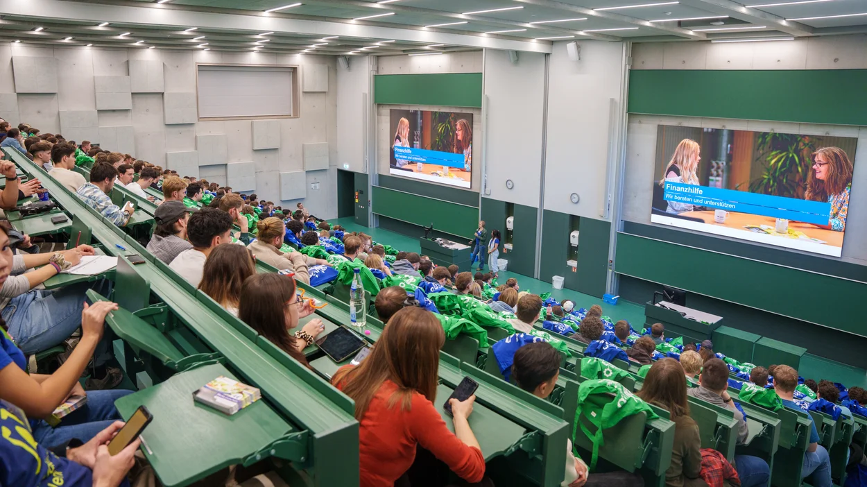 Hörsaal mit Studierenden, an der Wand ein Monitor mit dem Titel "Finanzhilfe"