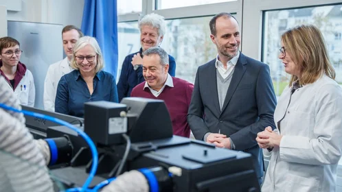 Eine Gruppe von Menschen steht an einem Gerät, das mit Schläuchen verbunden ist. Rechts im Bild erklärt eine Professorin im Labor etwas.