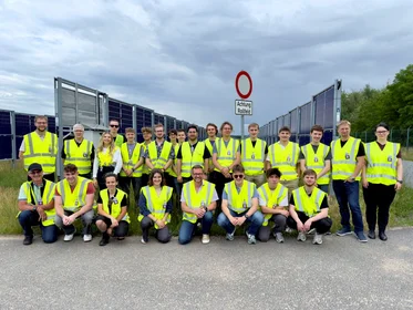 Eine Gruppe Studierender in leuchtend gelben Westen vor einem Schild "Achtung Rollfeld" und vertikalen Solarmodulen