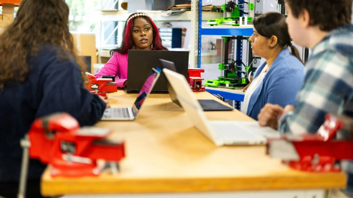 Gruppe in Werkraum bespricht sich, Fokus auf schwarzer Frau in pinkem Kostüm