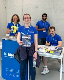 Drei Mitarbeitende in blauen THB-Shirts halten Flyer zum HIT und der Nacht der Wissenschaft in die Kamera