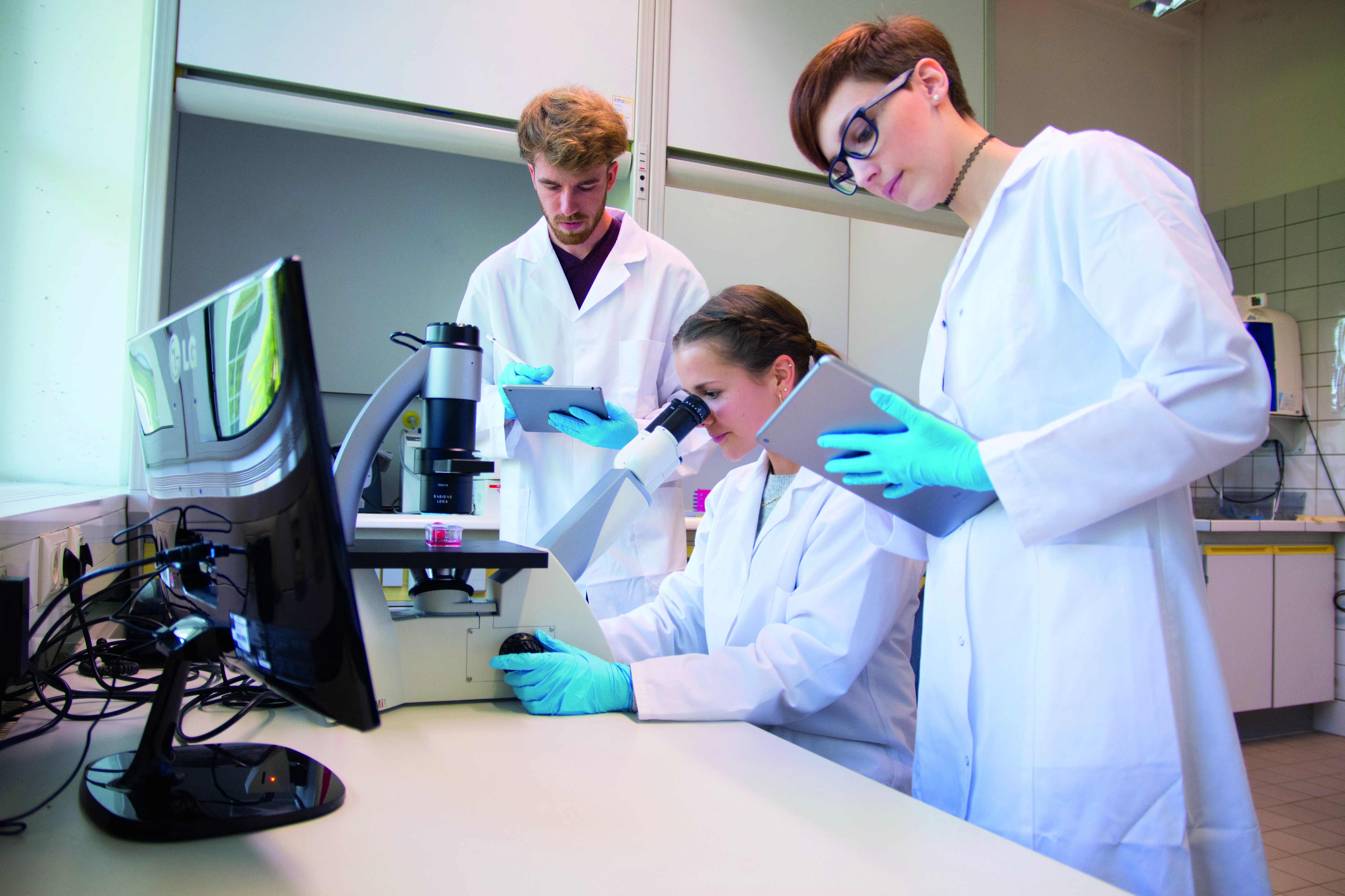 Das Bild zeigt drei Studierende im Labor, eine Studentin schaut in ein Mikroskop, die anderen stehen daneben und arbeiten mit. 