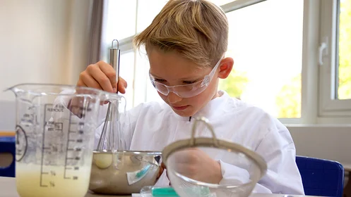 Das Bild zeigt einen Jungen im Laborkittel, der einen Schneebesen in einer Edelstahlschüssel rührt.