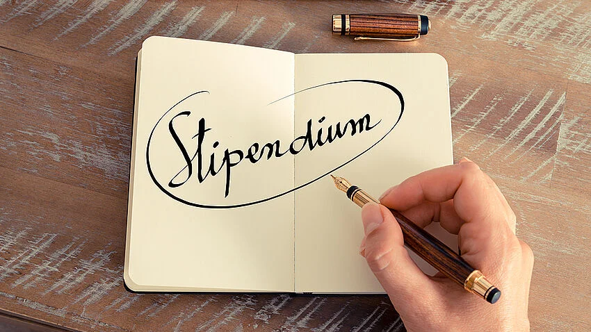 Aufgeschlagenes Notizbuch, eine Hand hält einen Füller, im Notizbuch steht "Stipendium"