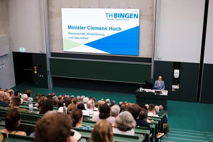 Mann hält Rede vor Publikum im Hörsaal, Präsentation im Hintergrund