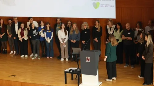 Große Gruppe Studierender und Betreuender stehen auf einer Bühne während eine Frau ins Mikrofon spricht