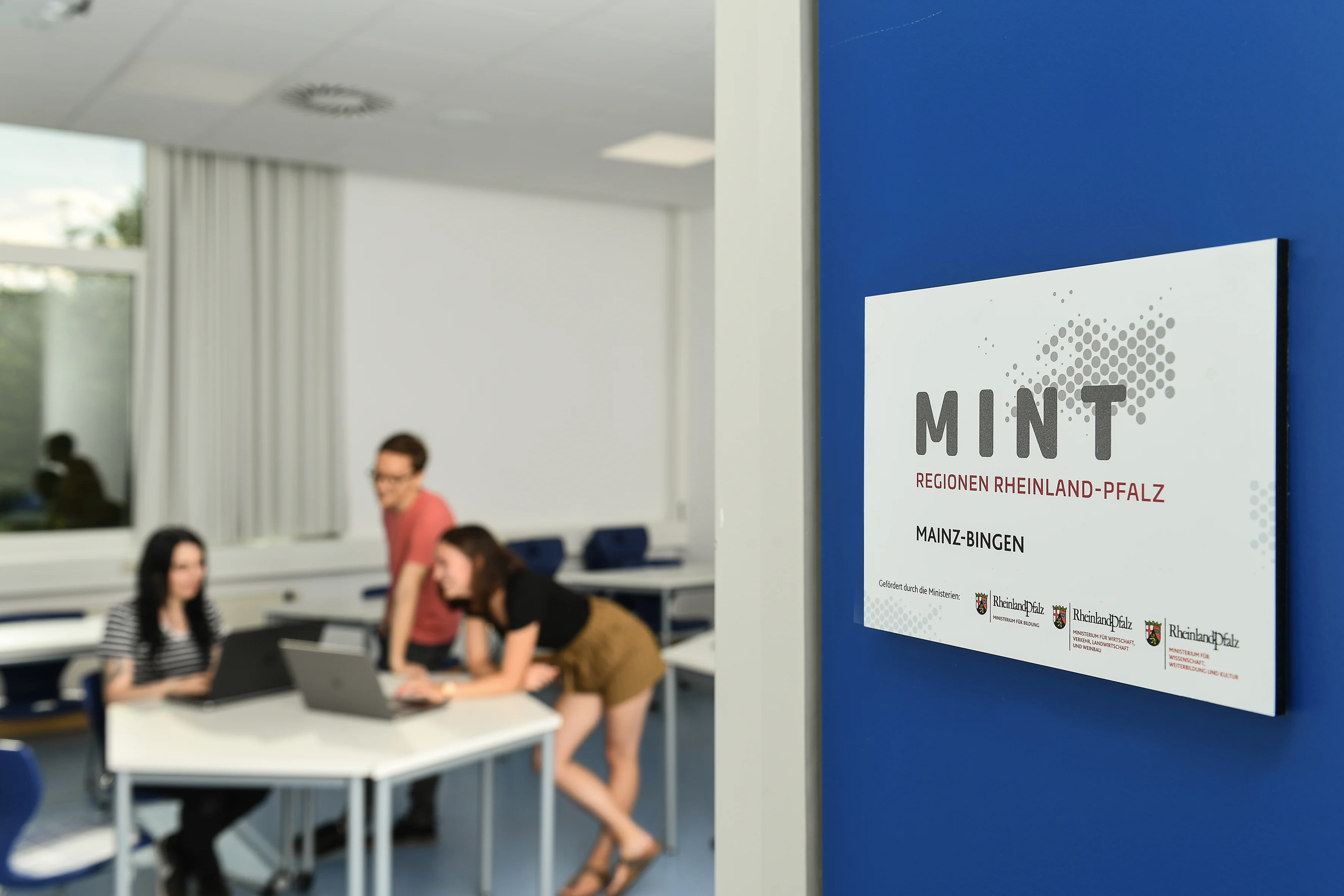 Weißes Schild MINT-Regionen auf blauer Tür, im Hintergrund junge Menschen