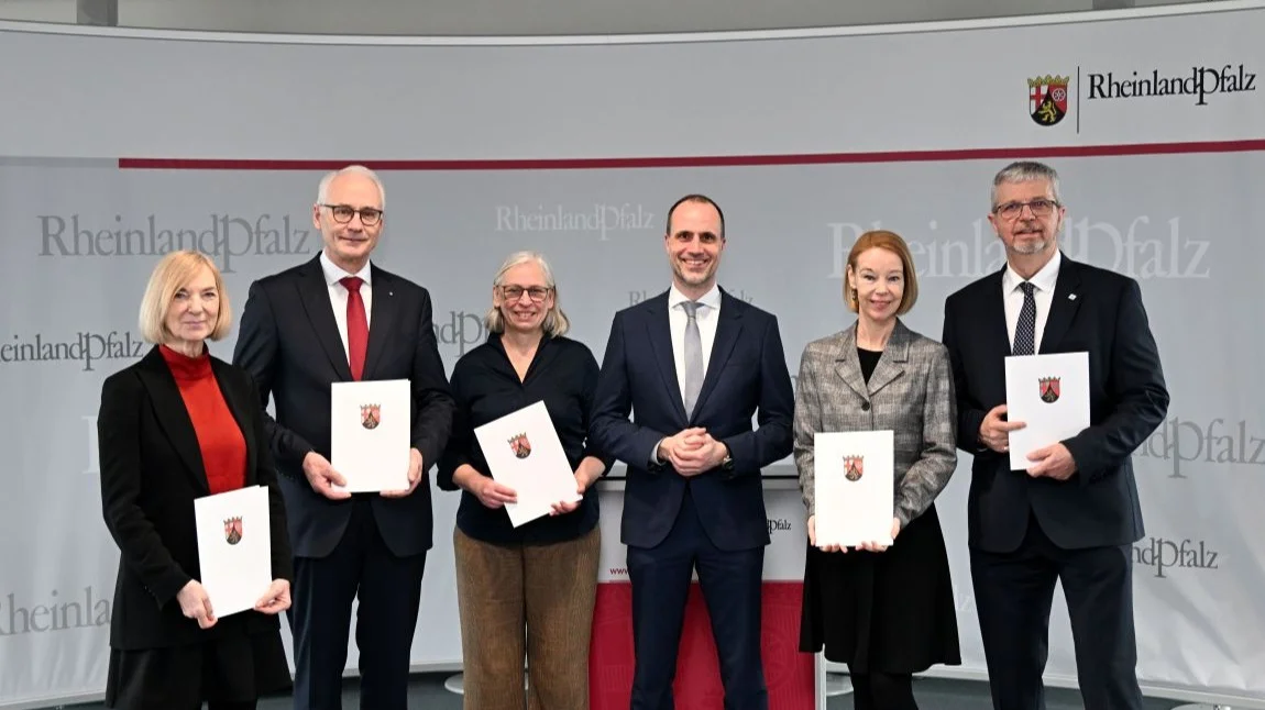 6 Personen (abwechselnd Frauen und Männer) stehen vor einer Wand mit Wappen des Landes Rheinland-Pfalz und halten Urkunden in den Händen