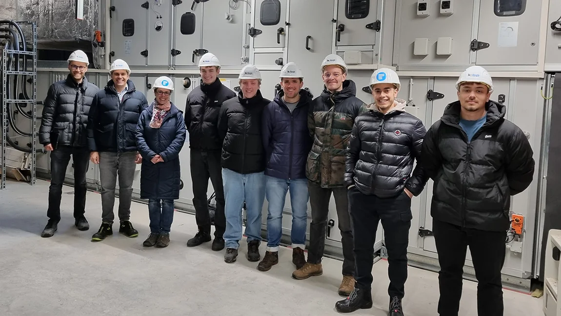 Gruppe Studierender mit Professorin vor einer Technikinstallation. Alle tragen weiße Helme