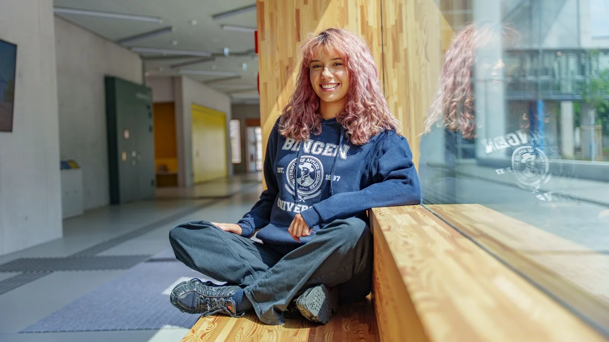Studentin mit welligen hellrosa Haaren und blauem TH-Hoodie sitzt locker am Fenster