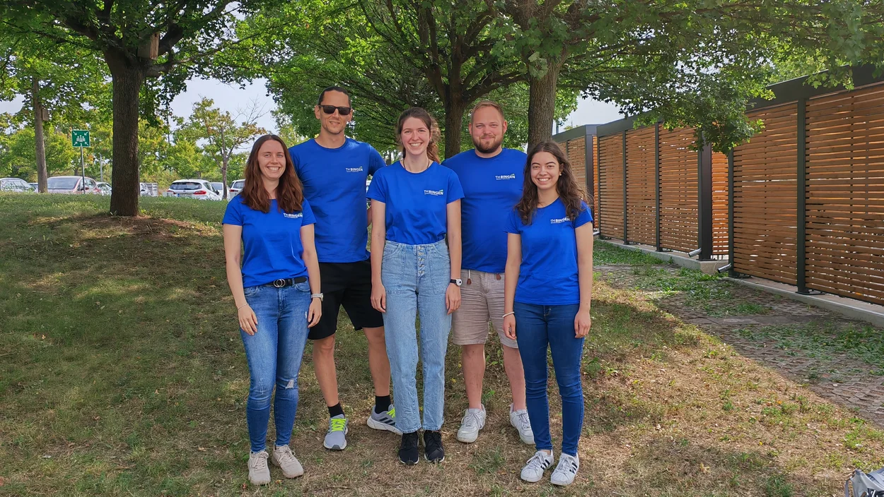 Fünf Studierende in blauen TH-Bingen-Shirts stehen im Grünen und lachen in die Kamera