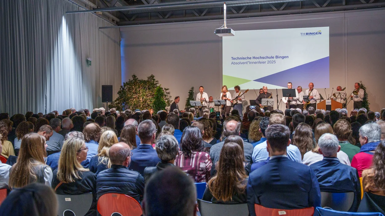 Menschen sitzen vor einer Bühne, auf der eine Band spielt. Im Hintergrund steht "Technische Hochschule Bingen Absolvent*innenfeier 2025"