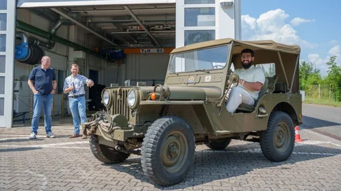 Geländewagen vor dem Fahrzeuglabor, ein dunkelhaariger Mann sitzt im Auto, zwei Männer im Hintergrund unterhalten sich lachend