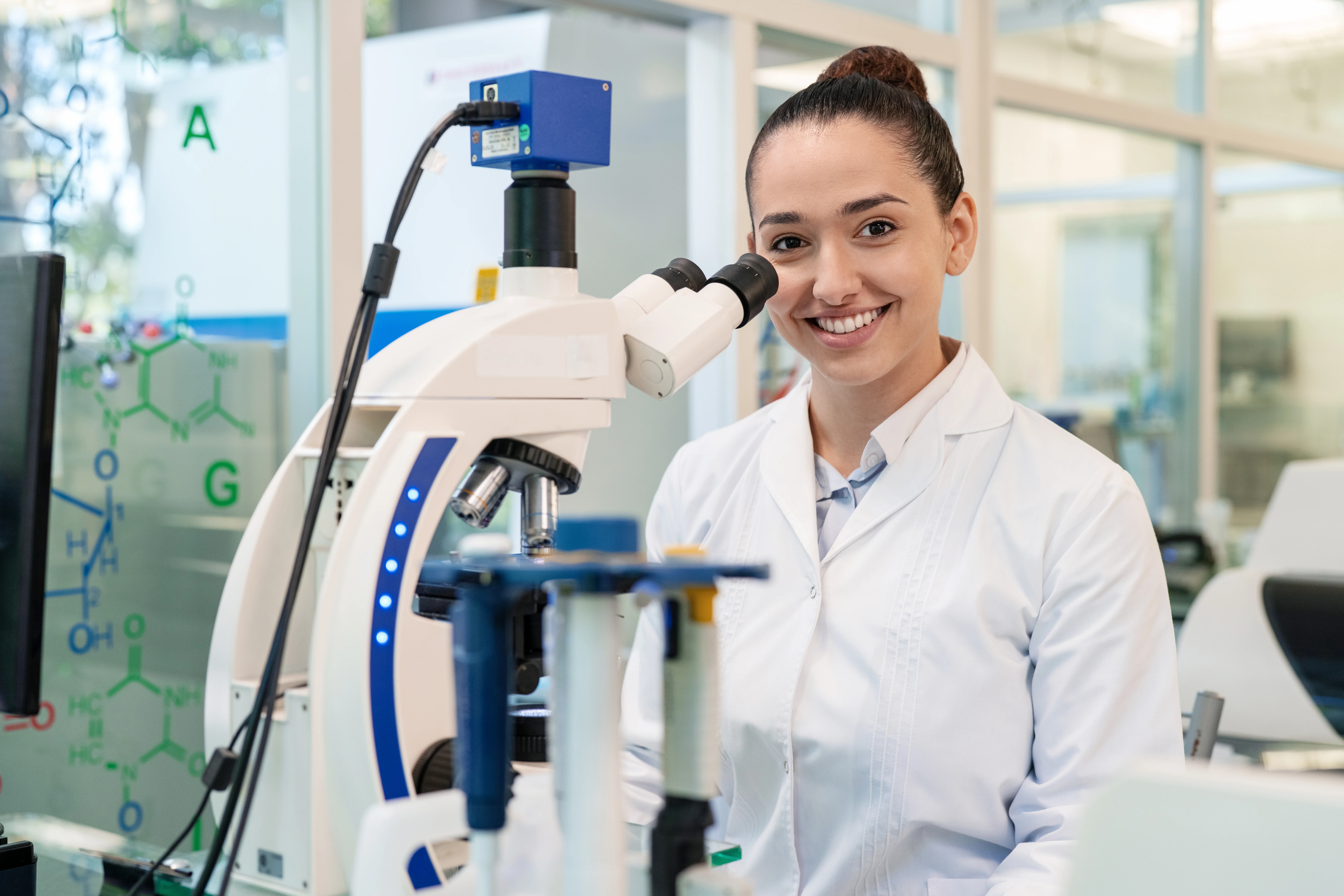 Porträt einer glücklichen Wissenschaftlerin, die am Mikroskop sitzt. Junge Ärztin, die im Labor arbeitet, schaut in die Kamera und lächelt.