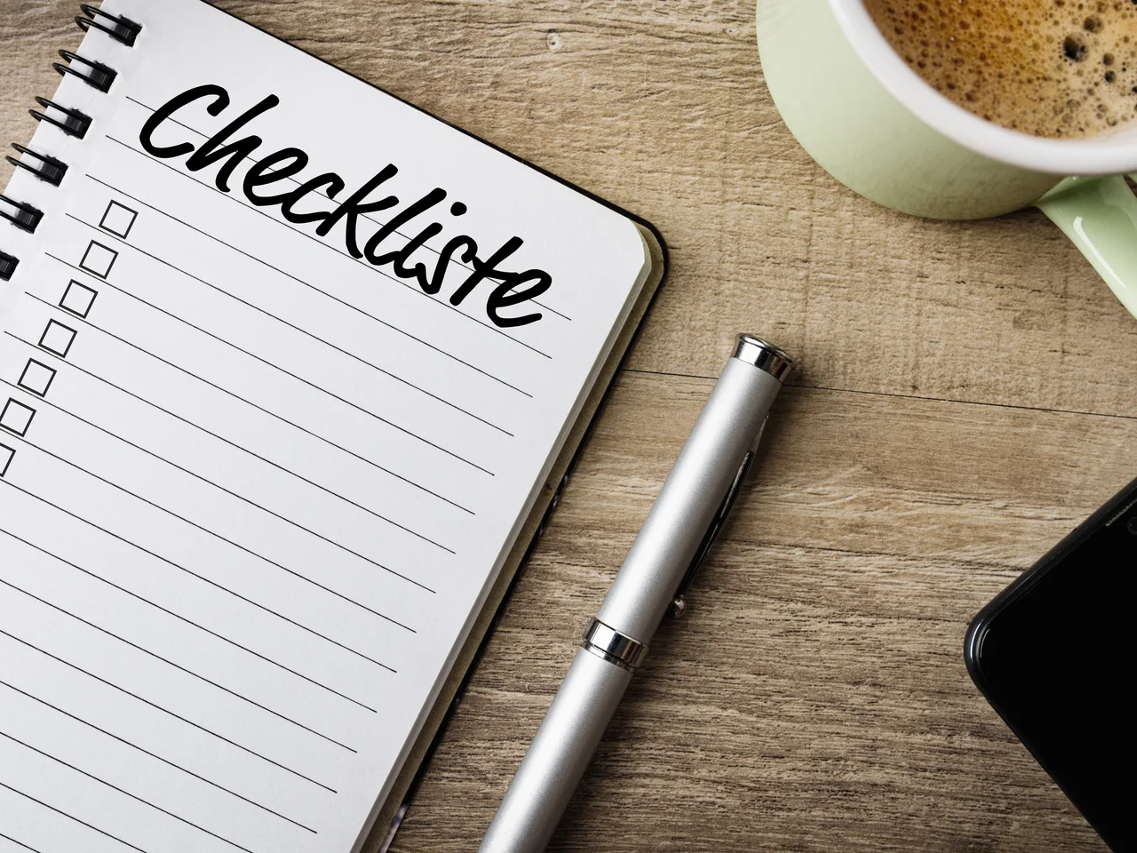 Eine Checkliste auf einem Holztisch, daneben ein Kugelschreiber und eine Kaffeetasse.