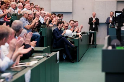 Publikum applaudiert im Hörsaal. Mann schaut lachend auf Bauhelm.