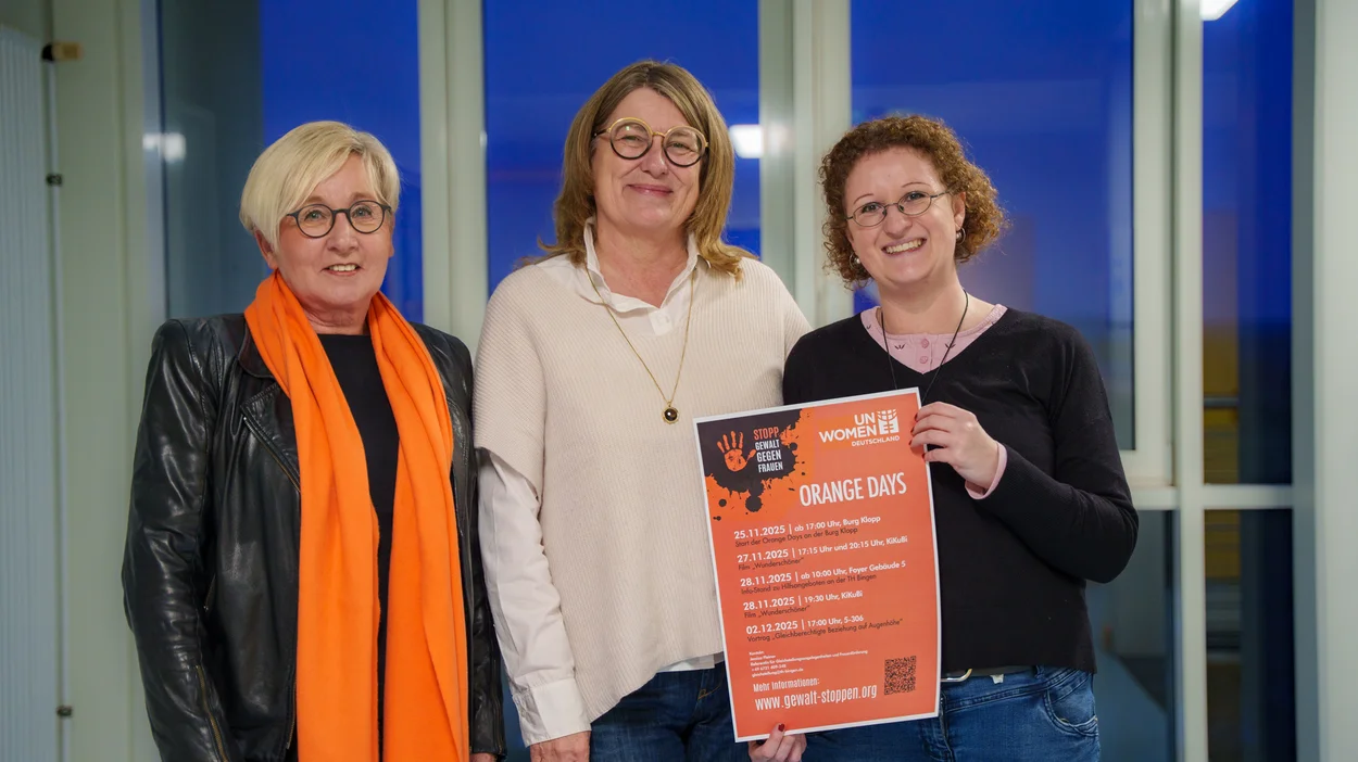 Eine Frau mit blonder Kurzhaarfrisur, orangenem Schal, eine Frau mit mittellangen Haaren und eine Frau mit mittellangen Locken, die ein orangefarbenes Veranstaltungsplakat hält