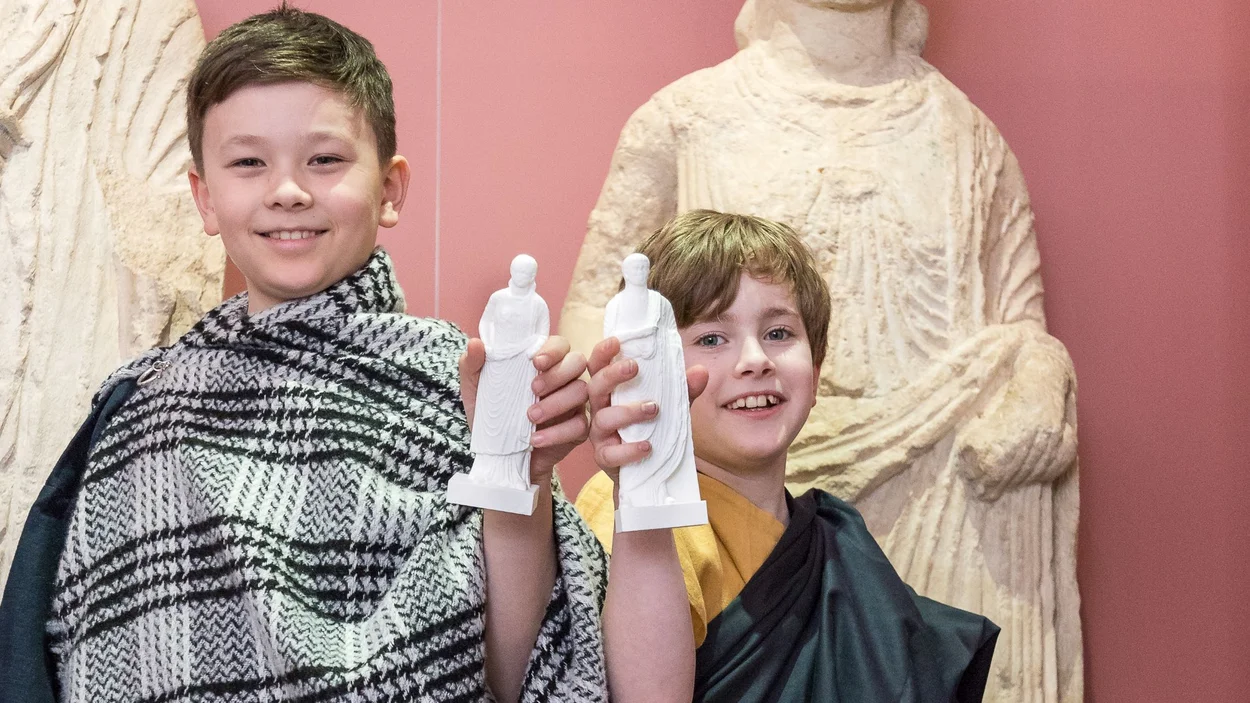 Zwei in römische Gewänder gekleidete Jungen halten selbst gegossene Figuren in die Kamera