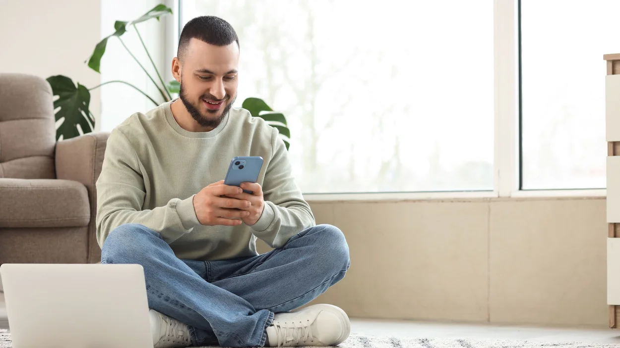 Student sitzt auf hellem Teppich auf dem Boden vor einer Couch und sucht etwas auf dem Handy