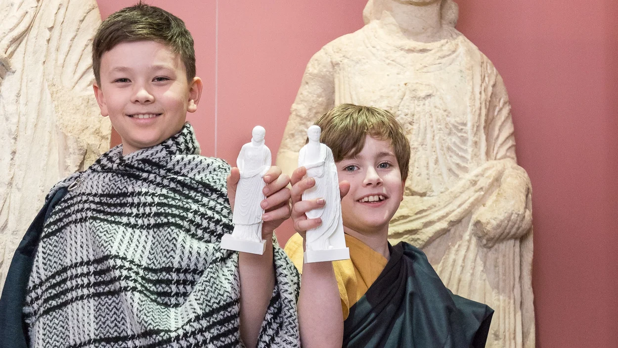 Zwei Jungen in Togakleidung halten selbst gegossene Römerfiguren in die Kamera
